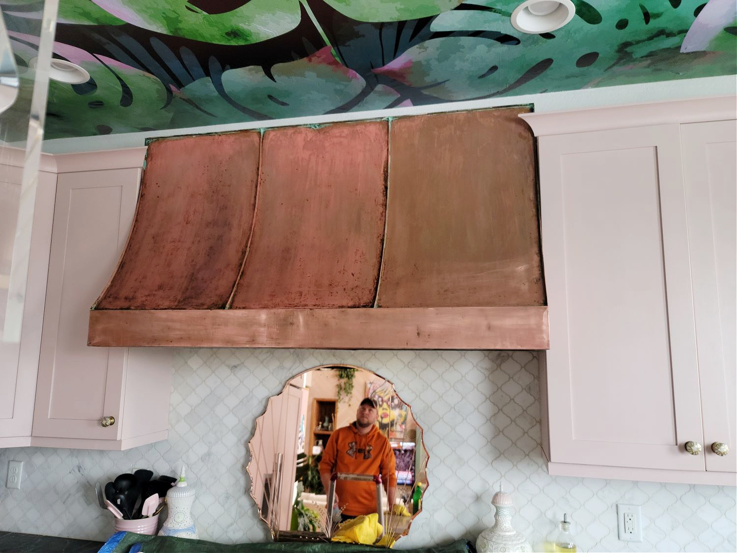 Copper Hood by popular CO home design blog, Eclectic Twist: image of a copper hood hanging above a blue gas range stove in a kitchen with pink cabinets, black and white marble countertop, white tile backsplash, and pendant lighting. 