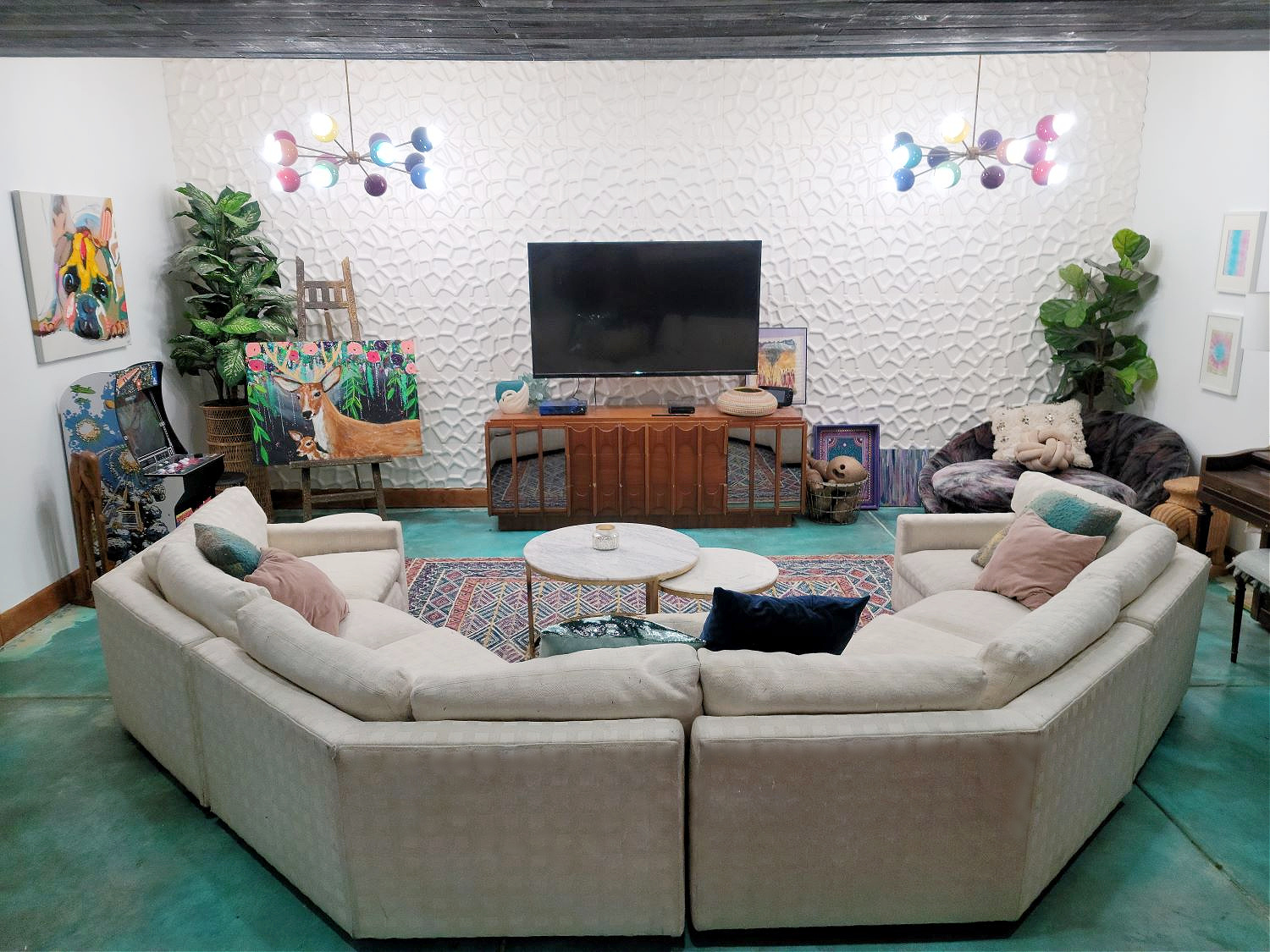 Basement Living Room by popular CO home design blog, Eclectic Twist: image of a basement living room with a multi color geometric area rug, white sectional couch, multi color mid century modern lights, red area rug, piano, colorful artwork, bar table and bar stools, indoor plants, flat screen t.v. and blue cement floors. 