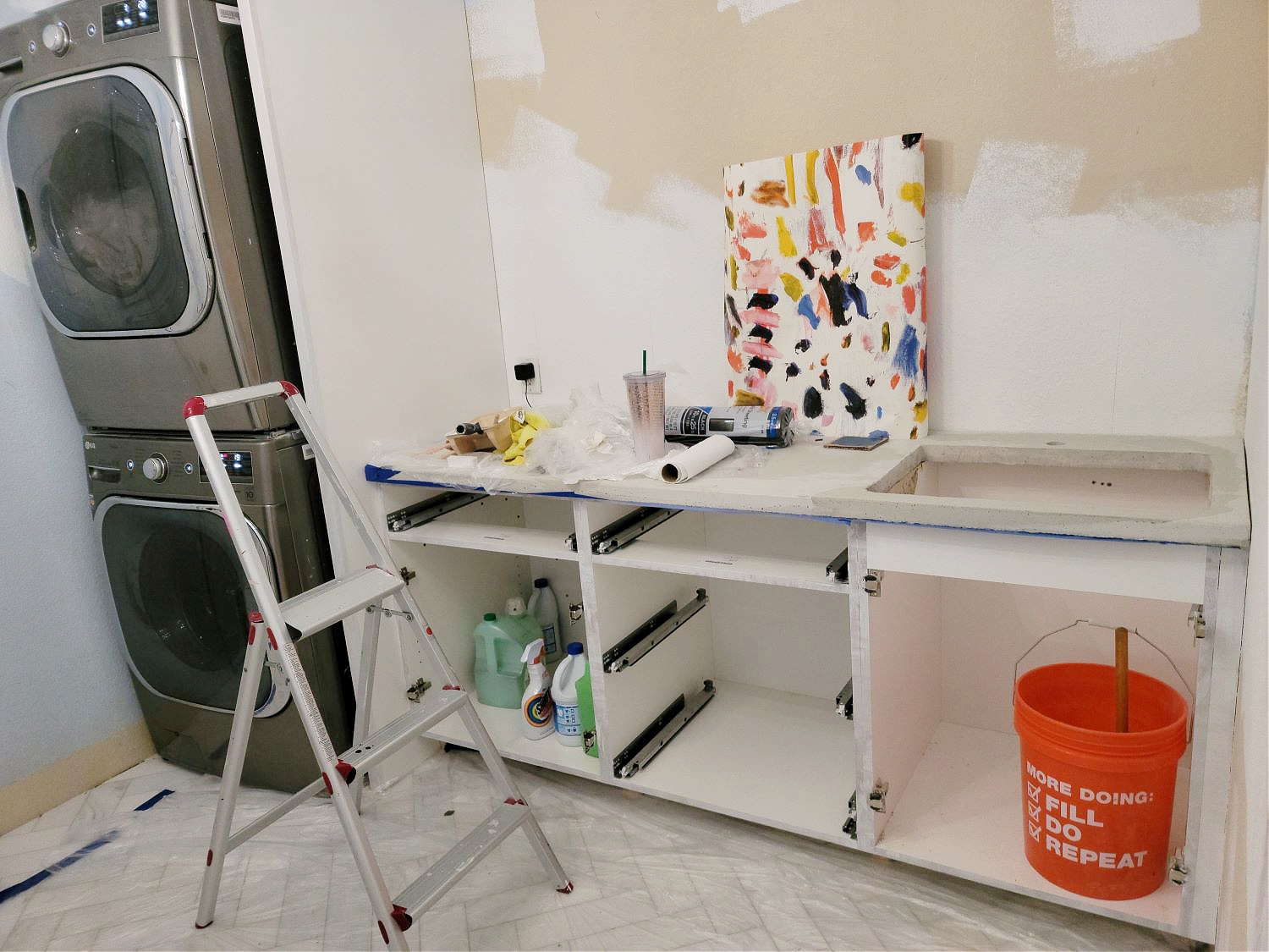 Mixing Your Own Paint Color by popular CO Home Design blog, Eclectic Twist: image of a remodeled laundry room with a stacked washer and dryer, unfinished cabinets, sheetrock, white marble chevron pattern tile flooring, metal step ladder, and a canvas with multi color paint smears on it. 