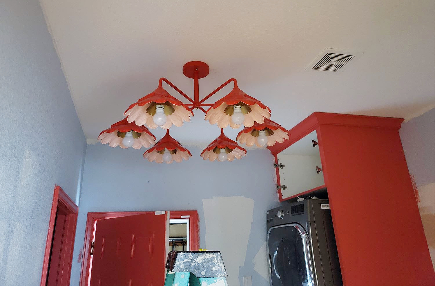Mixing Your Own Paint Color by popular CO Home Design blog, Eclectic Twist: image of red metal chandelier hanging from the ceiling of a laundry room with a stacked washer and dryer and coral pink cabinets and door. 